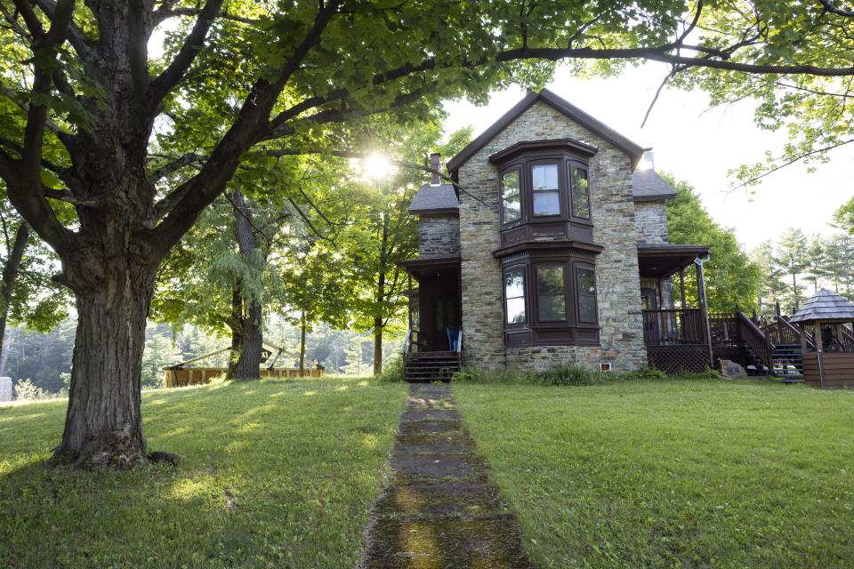 A stone house sits on a green lawn at the end of a long sidewalk, surrounded by trees.