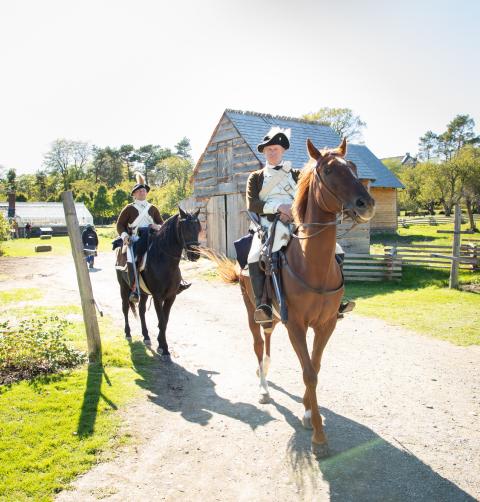 Two men in colonial clothing ride horses through a vineyard.