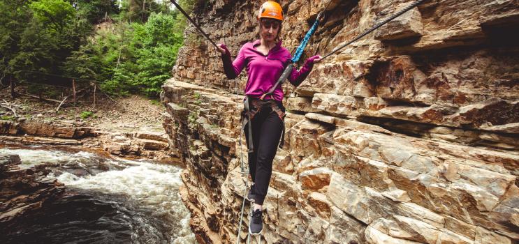 A woman walks across a metal wire on a ropes course in a narrow, rocky gorge