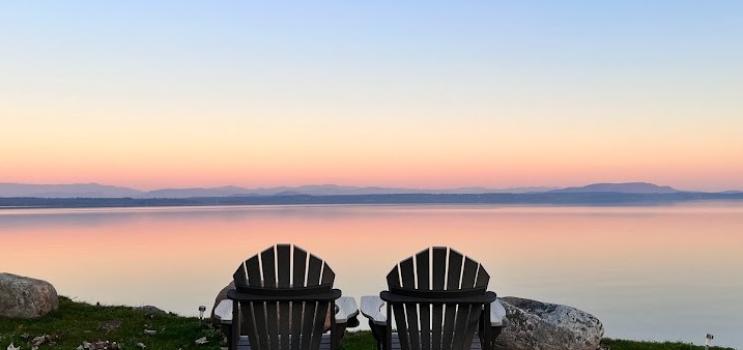 Two Adirondack chairs sit by a quiet lakeshore during a pink- and blue-hued summer sunset. 