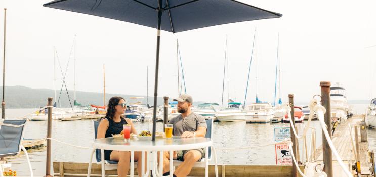 A man and woman sit at an umbrella-shaded table at a lakeside marina restaurant, with lake and sailboats in the background.