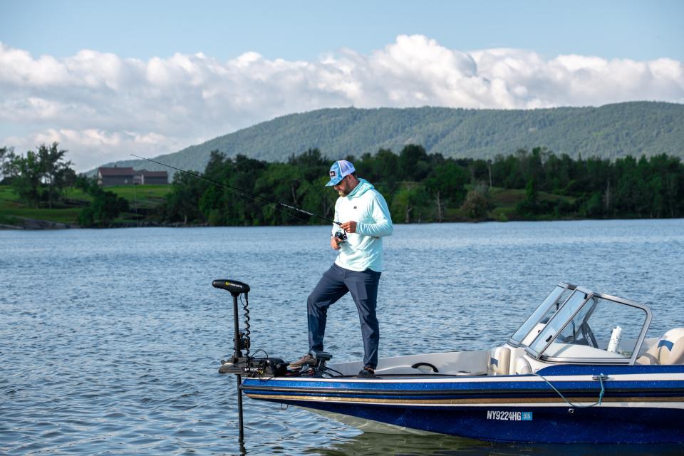 A man reels in a fish on the edge of a boat with farms in the background.