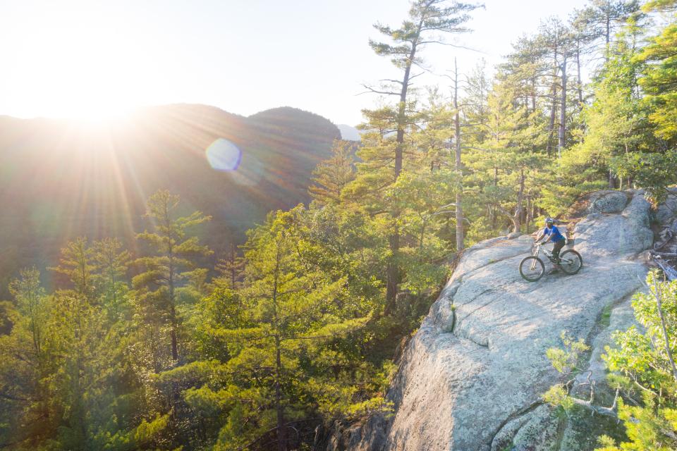 A man sits on his bike on a rocky cliff surrounded by forests and mountains.
