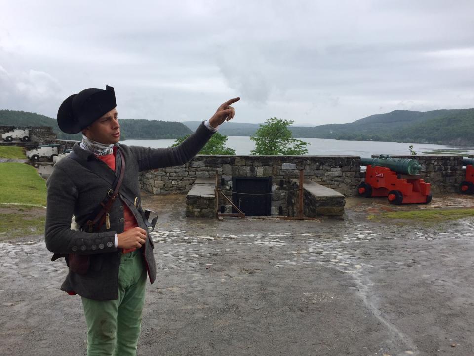 A Fort Ticonderoga war reenactor stands in front of an outdoor landscape, pointing off into the distance.