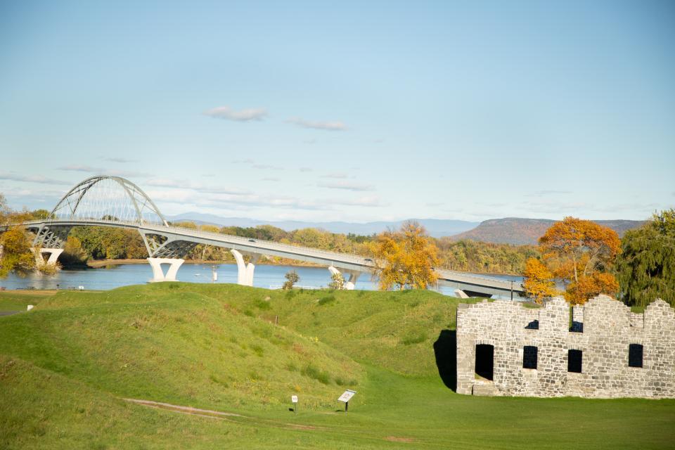 Stone ruins of an early colonial American fort look out over orange-leaved trees, a bridge, and lake.