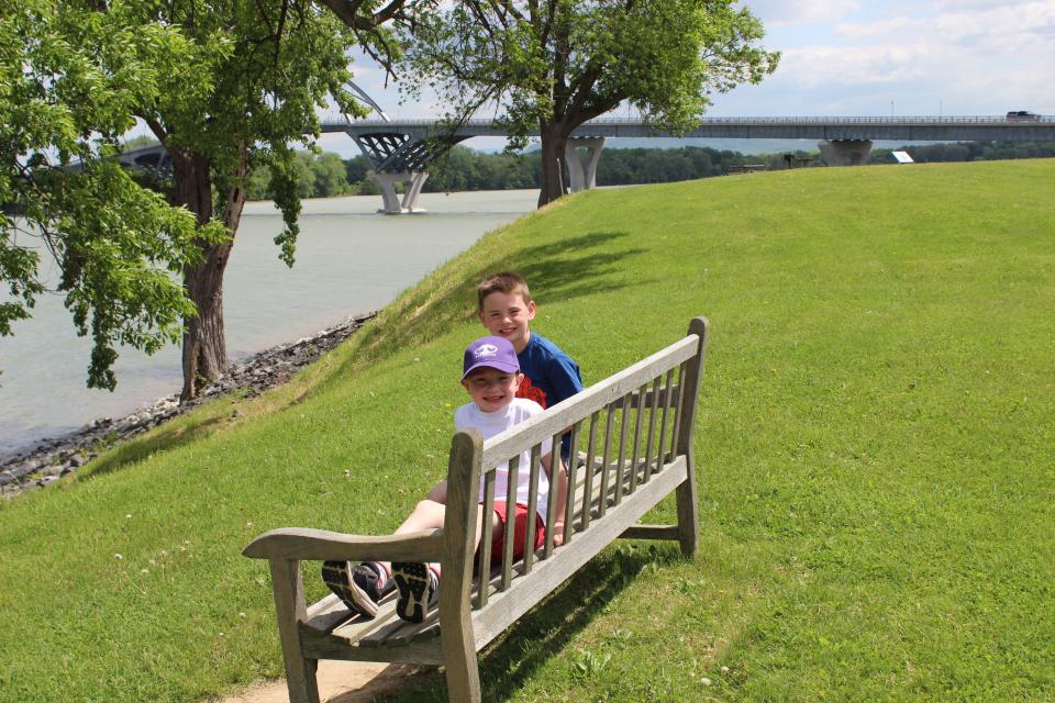 Two young boys smile for a picture on a park bench overlooking the water.