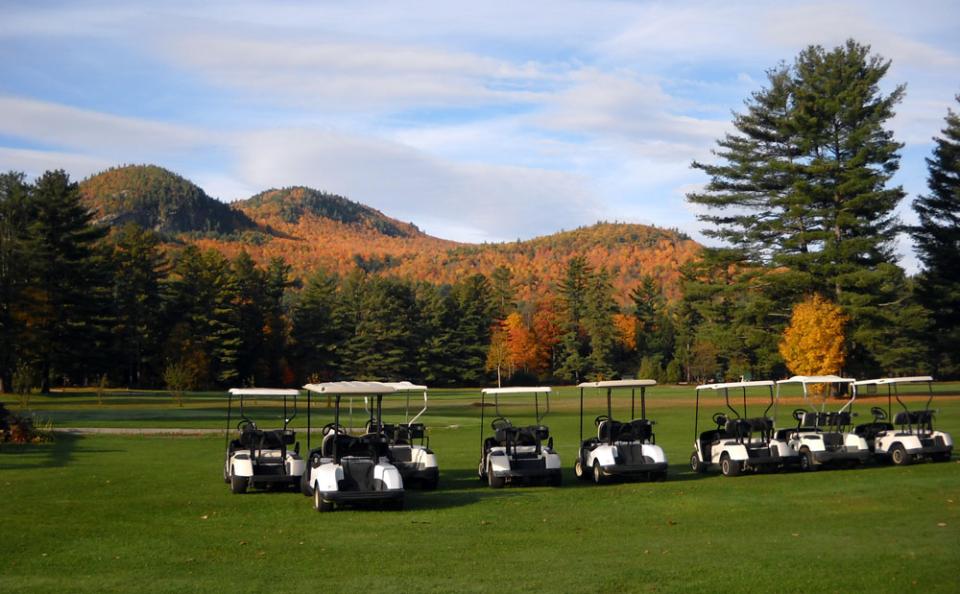 Golf carts lined up in front of a green course; low mountains with fall foliage are in the background.