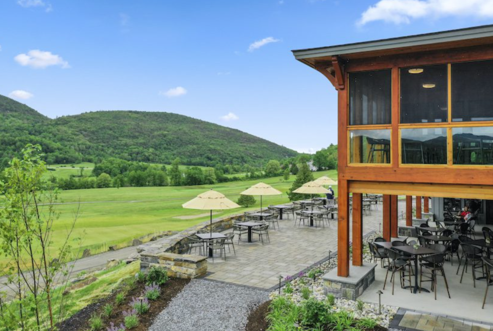 A two-story golf club and patio with tables and umbrellas look out over a bright green golf course, with mountains in the background.