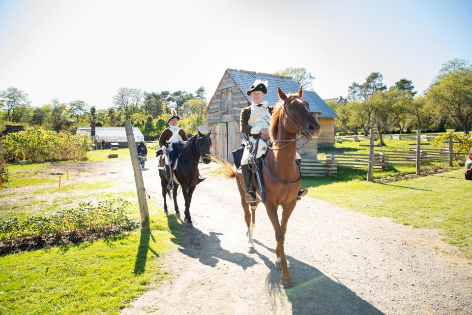 Colonial reanactors ride horses down a dirt road.